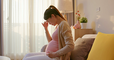  Schwangere Frau mit Kopfschmerzen sitzt auf dem Sofa, hält ihren Bauch und lehnt sich zurück, im sonnigen Zimmer mit weichen Farben.